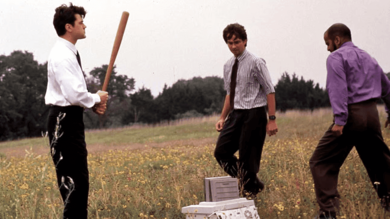 Three men in dress shirts and ties stand in a grassy field; the man on the left raises a wooden baseball bat over a battered office printer on the ground between them — the printer-destruction scene from the film Office Space.